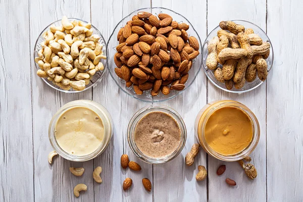 Modern refah ve veganlık kavramı. Beyaz ahşap bir masada kavanozlar dolusu badem, fıstık ezmesi ve fıstık ezmesi. Sağlıklı bir kahvaltı için organik, ev yapımı fıstık ezmesi..