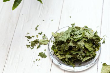 Beyaz ahşap bir masada cam bir tabakta kurutulmuş üzümlü yapraklar. Organik, doğal, kuru bitki çayıyla kompozisyon