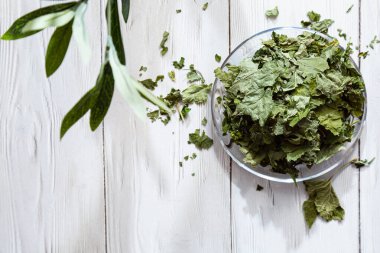 Beyaz ahşap bir masada cam bir tabakta kurutulmuş üzümlü yapraklar. Organik, doğal, kuru bitki çayıyla kompozisyon