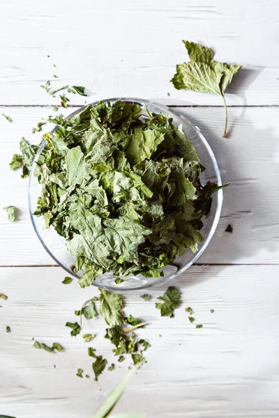 Beyaz ahşap bir masada cam bir tabakta kurutulmuş üzümlü yapraklar. Organik, doğal, kuru bitki çayıyla kompozisyon