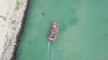 Turkuaz denizde kıyı boyunca yelken açan kırmızı bir balıkçı teknesinin kuş bakışı görüntüsü. Okyanusta seyreden yerel bir geminin drone görüntüsü.