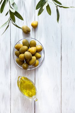 Beyaz ahşap bir masada zeytinyağı ve bir kase yeşil zeytin. Modern, durgun yaşam, zeytinli cam kase, zeytin dalı ve kopyalama alanı..