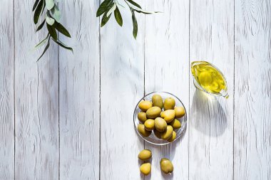 Beyaz ahşap bir masada zeytinyağı ve bir kase yeşil zeytin. Modern, durgun yaşam, zeytinli cam kase, zeytin dalı ve kopyalama alanı..