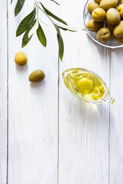 Beyaz ahşap bir masada zeytinyağı ve bir kase yeşil zeytin. Modern, durgun yaşam, zeytinli cam kase, zeytin dalı ve kopyalama alanı..