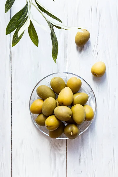 A bowl of green olives. Minimal still life of an olive branch and a glass bowl of olives on a white wooden table. Copy space
