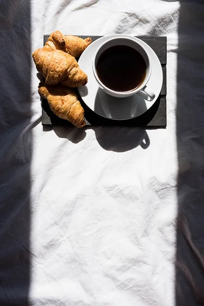 朝の日差しの中でベッドの上でコーヒーカップとクロワッサン 興味深い影とコピースペースとトレンディな静物画 — ストック写真