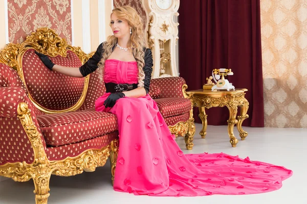 Stylish blonde in long red dress is sitting on luxurious  sofa — Stock Photo, Image