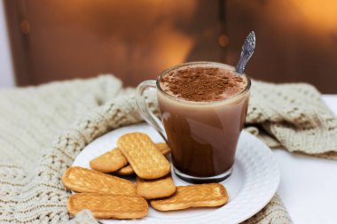 Beyaz bir masada beyaz bir tabakta bir fincan kakao ve kurabiye ve örülmüş bir eşarp.