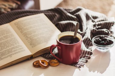 Kırmızı bardakta bir fincan sıcak kahve, kurabiyeler ve beyaz ahşap bir masada eski bir kitap.