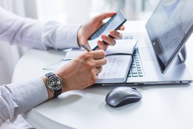 Bir masada oturan bir adamın resmi, bir elinde telefon, diğer elinde tükenmez kalem, bir deftere bir bilgisayar faresinin yanında yazan bir adam.