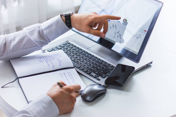 The image of the hands of a young man sitting at a table writing in a Notepad with a ballpoint pen, the second hand shows the laptop screen. On the screen of the laptop depicts graphs