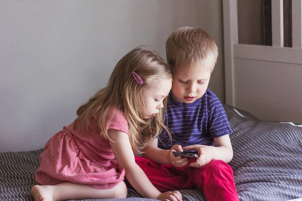 A blond boy and a girl sit on the bed and look at the smartphone. Online communication, video communication, education