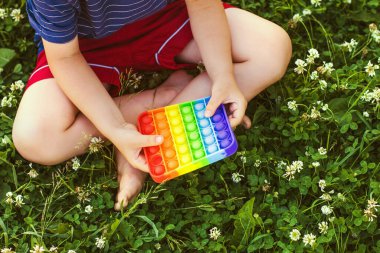 Yeşil çimlerin üzerinde oturan kişiliksiz küçük çocuk Pop It ile oynuyor. Popo kabarcığı esnetilebilir hareketli duyusal oyuncak boşaltımı sağlar