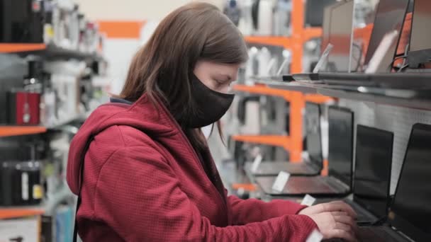 Een vrouw met een masker tegen het virus koopt een computer. — Stockvideo