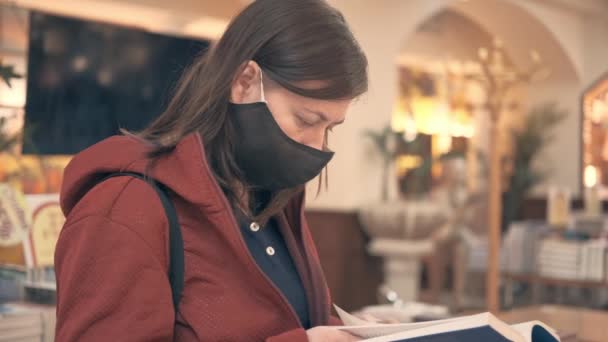 Chica con máscara médica elige un libro de papel en una tienda — Vídeos de Stock