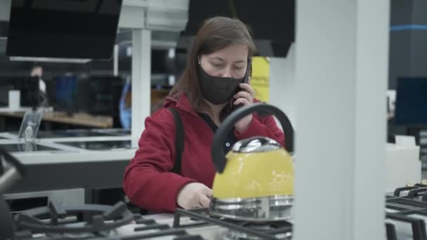 Женщина в маске выбирает новую плиту в магазине бытовой техники — стоковое видео