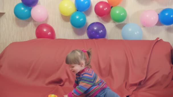 Маленькая забавная девочка играет с игрушкой-погремушкой. Вокруг надувного, воздушные шары — стоковое видео