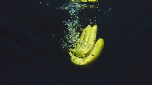 Bando maduro de bananas amarelas no fundo azul cai lentamente na água — Vídeo de Stock