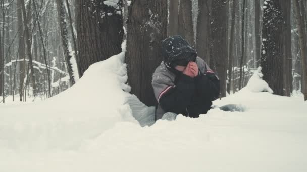 Mannen bär solglasögon sitter midjan djupt i snön bredvid trädet och fryser — Stockvideo