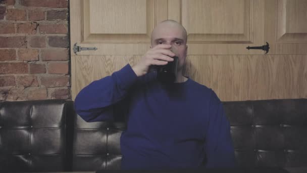 Белый мужчина с бородой сидит в темном пабе и пьет тёмное пиво из стакана. — стоковое видео