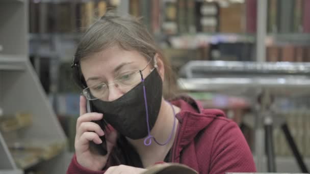 Kvinna i mask och glasögon bläddrar igenom boken i biblioteket och pratar i telefon — Stockvideo