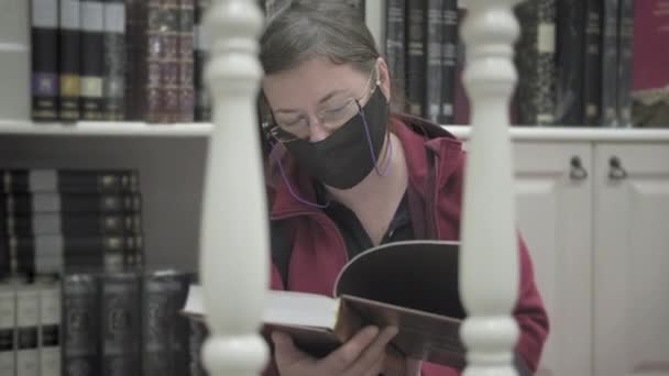 Frau mit Brille und Maske gegen Virus sitzt auf Bibliotheksboden und liest Buch — Stockvideo