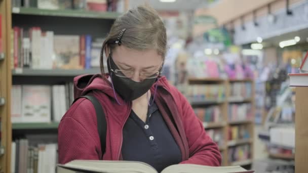 Mujer caucásica en gafas y máscara contra el virus lee en la biblioteca y piensa — Vídeos de Stock