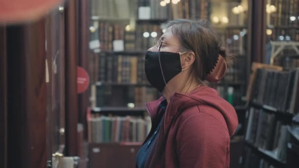 Женщина в очках и маске против вируса выбирает книгу для покупки в антикварном магазине — стоковое видео