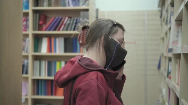 Nő ellen vírus maszk és szemüveg áll a könyvtárban, és beszél telefonon — Stock videók