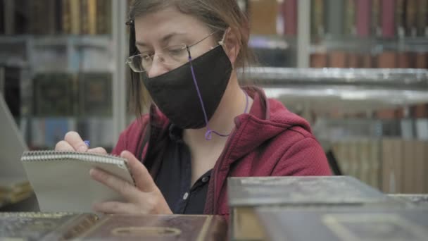 Kobieta w okularach i masce w księgarni pisze tytuły książek do notatnika — Wideo stockowe