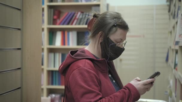 Mulher usando óculos e máscara na livraria. Ela escreve títulos livros em telefone — Vídeo de Stock