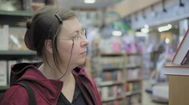 Zincirli gözlüklü ciddi bir beyaz kadın kitapçıdan kitap alır.