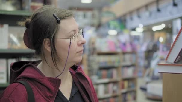 Serieuze blanke vrouw met bril op ketting koopt een boek in boekwinkel — Stockvideo