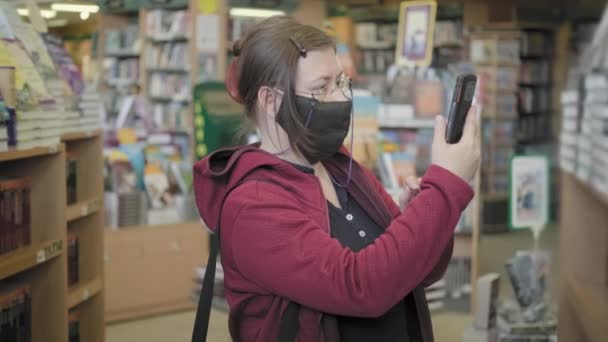 Kobieta w okularach i masce w bibliotece robi zdjęcia książek na telefonie — Wideo stockowe