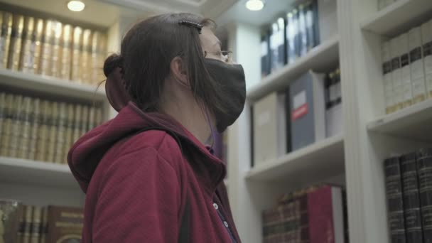 Mulher usando óculos e máscara contra vírus escolhe livro para comprar na livraria — Vídeo de Stock