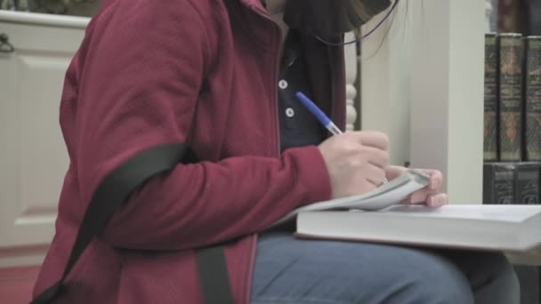 Женщина в очках и маске сидит на ступеньках библиотеки, читает книги, делает заметки — стоковое видео