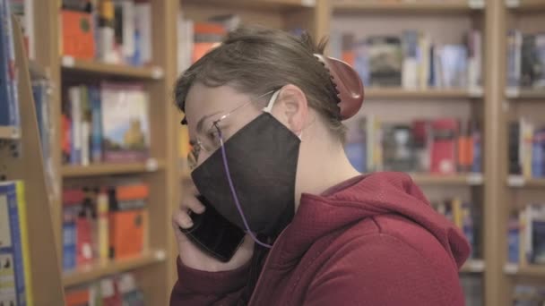 Woman in against virus mask and glasses stands in book store and talks on phone — Stock Video