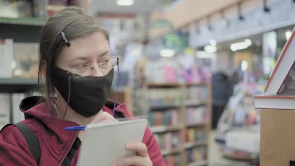 Vrouw met bril en masker schrijft namen van boeken op in de winkel in een notitieboekje — Stockvideo