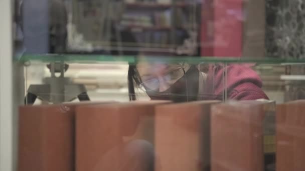 Donna che indossa occhiali e maschera contro il virus sceglie il libro da acquistare in libreria — Video Stock