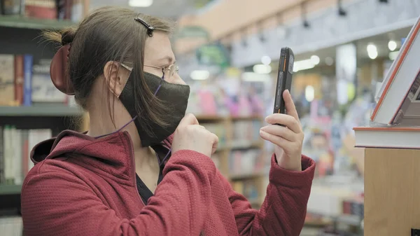 Woman wearing glasses and mask in bookstore takes pictures of books on her phone — Stock Photo, Image