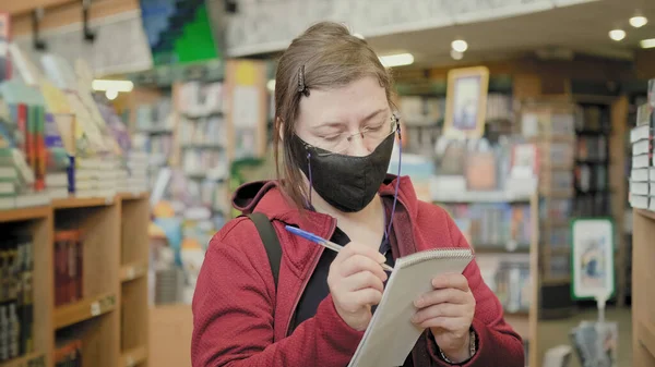 Woman in glasses and mask against virus in bookshope makes notes in notebook — Stock Photo, Image