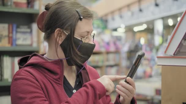 Mulher de óculos e máscara contra vírus na livraria. Ela digita na tela do telefone — Vídeo de Stock
