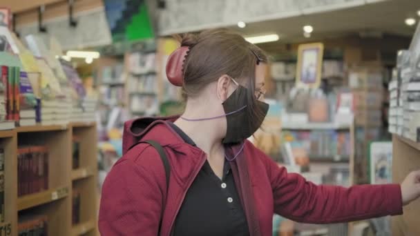 Woman wearing glasses and mask against virus chooses book in library — Stock Video