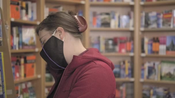 Caucasian woman wearing glasses and a mask against virus buys a book in a store — Vídeo de Stock