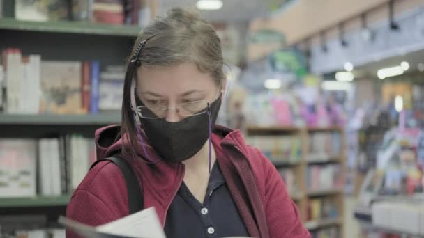 Woman wearing glasses and a mask against virus flips through a book in the store — Wideo stockowe