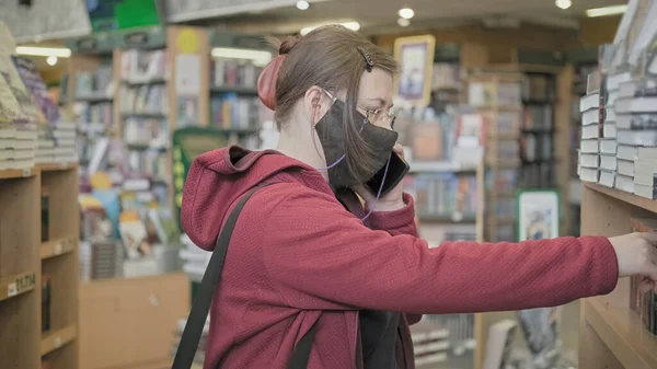 Virüs maskesi ve gözlüğe karşı olan kadın kitapçıda duruyor ve telefonla konuşuyor. — Stok fotoğraf