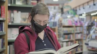 Gözlüklü ve maskeli ciddi bir beyaz kadın kütüphanede virüs kitapları okuyor.