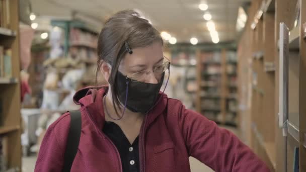 Kobieta w okularach i masce przeciw wirusowi bierze książkę z półki księgarni — Wideo stockowe