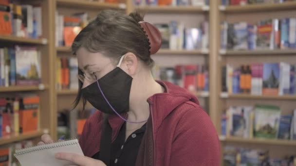 Библиотекарь в очках и маске составляет список книг на полке читального зала — стоковое видео