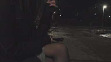 Uzun siyah saçlı mini etekli kız bankta oturur ve sigara içer.